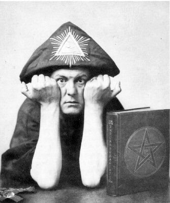 Aleister Crowley wearing a triangular all-seeing-eye hat beside a book bearing a pentagram.