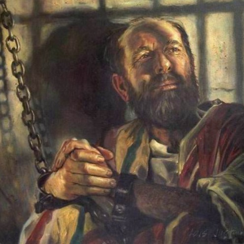 A man in shackles looks upward toward light in a prison cell.